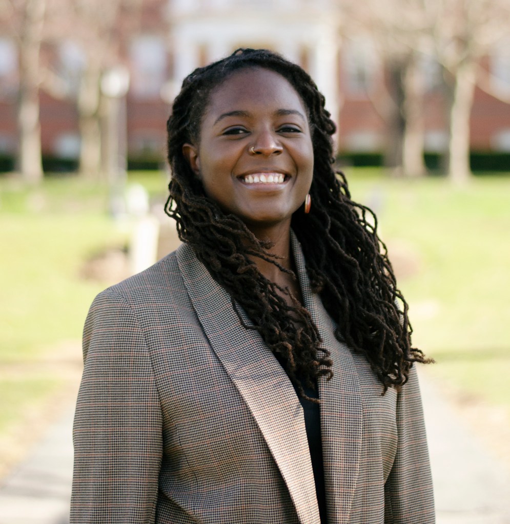 A photo of Aja Kennedy standing outside. She is wearing a grey blazer, smiling, and looking into the camera.